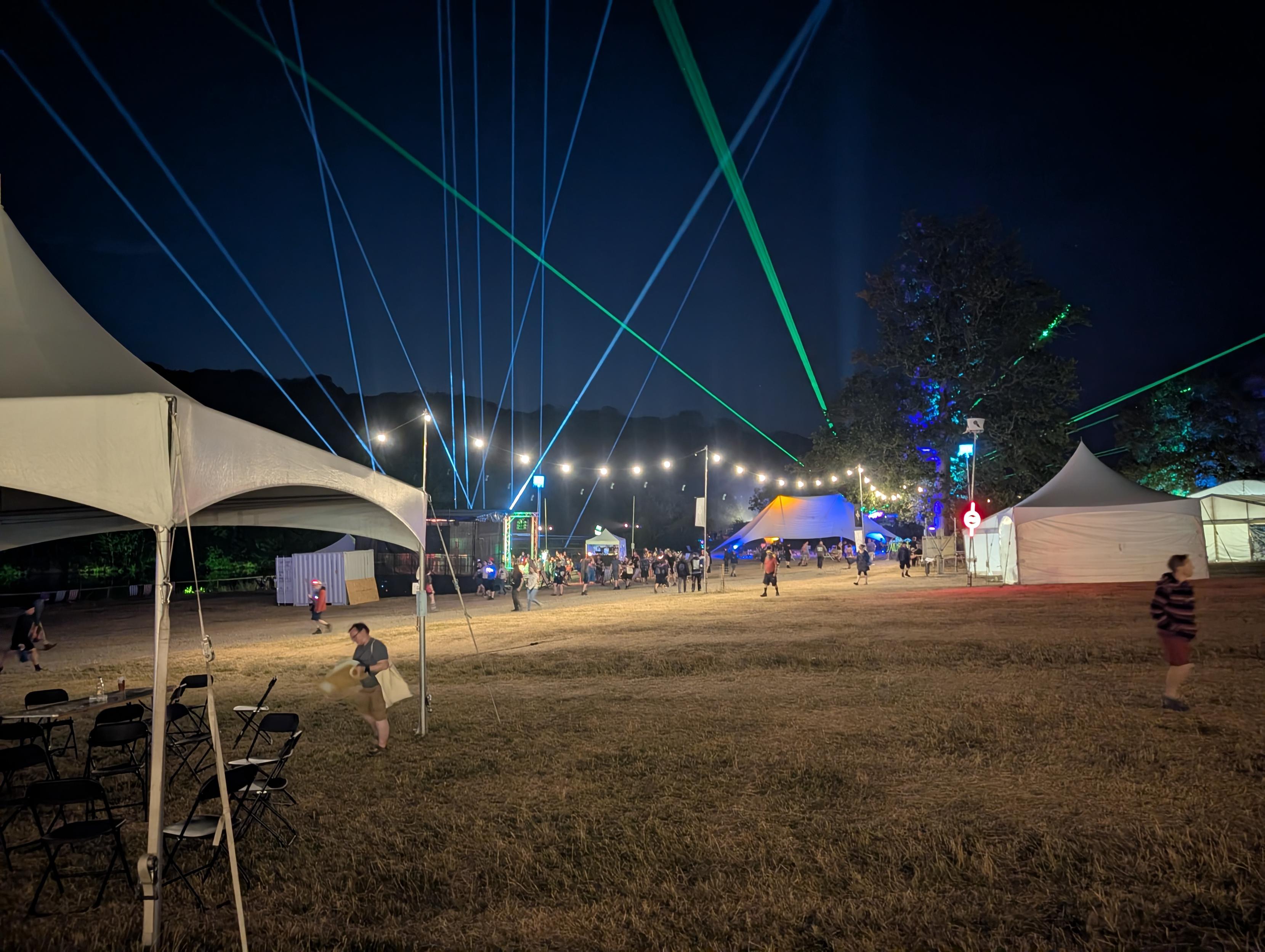 A field containing tents at night. It is well lit and the sky is full of spotlights and green lasers.