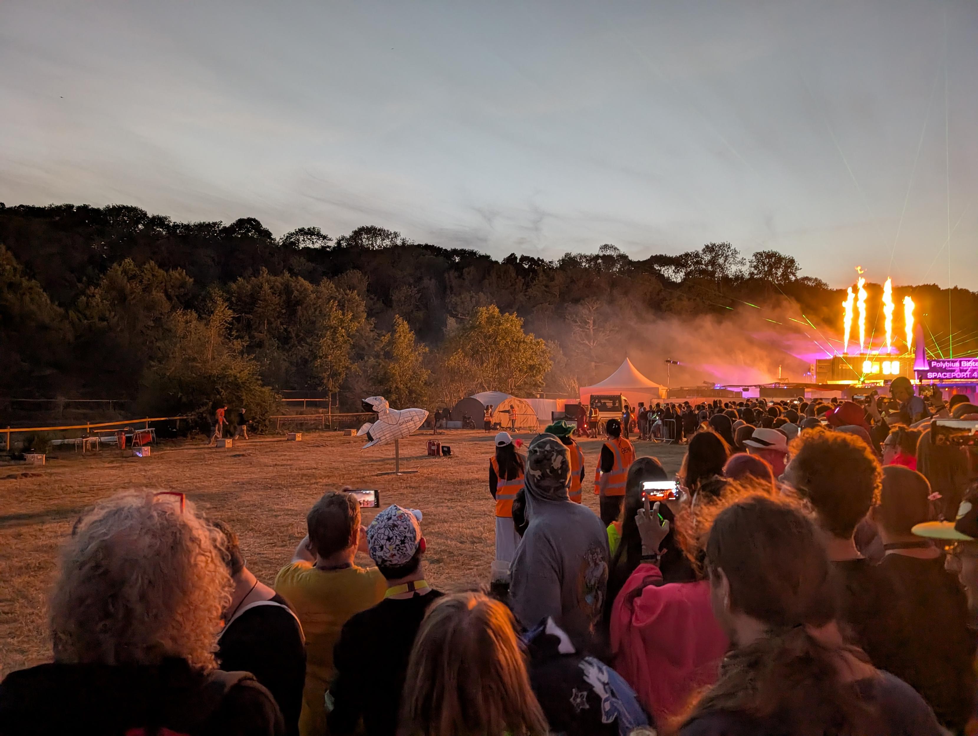 A field containing an effigy of Elon Musk riding a rocket. Flames rise in the background as the Null Sector stage kicks off. There is a large crowd.